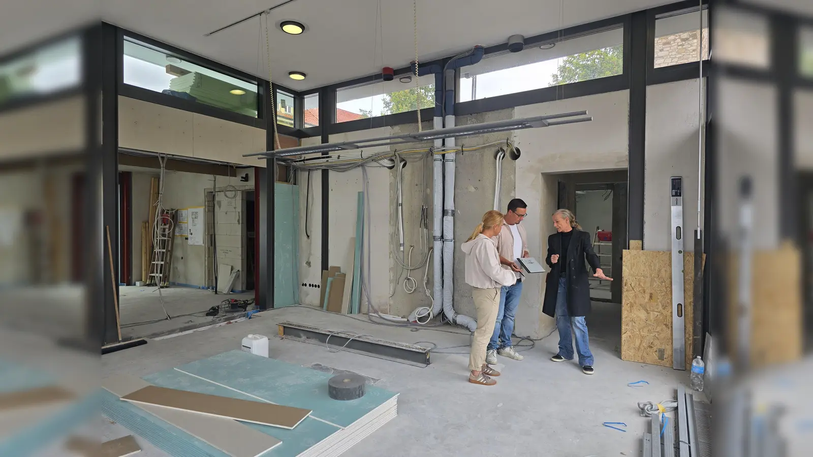 Das Herzstück des Marktplatzcafés wird die Holzrückwand im Gastraum sein. Sie wird sich künftig dort befinden, wo jetzt noch Leitungen und Rohre frei auf der Wand liegen. Silke Bergerhoff (rechts) und Katrin Micheel (links) von middel Büroeinrichtungen GmbH &amp; Co. KG planen in enger Abstimmung mit Pächter Walter Frühauf die Einbaumöbel des neuen Cafés. Die Holzrückwand ist mit speziellen Akustik-Elementen ausgestattet und wird zusätzlich zur Wohlfühlatmosphäre beitragen. (Foto: Stadt Warburg)