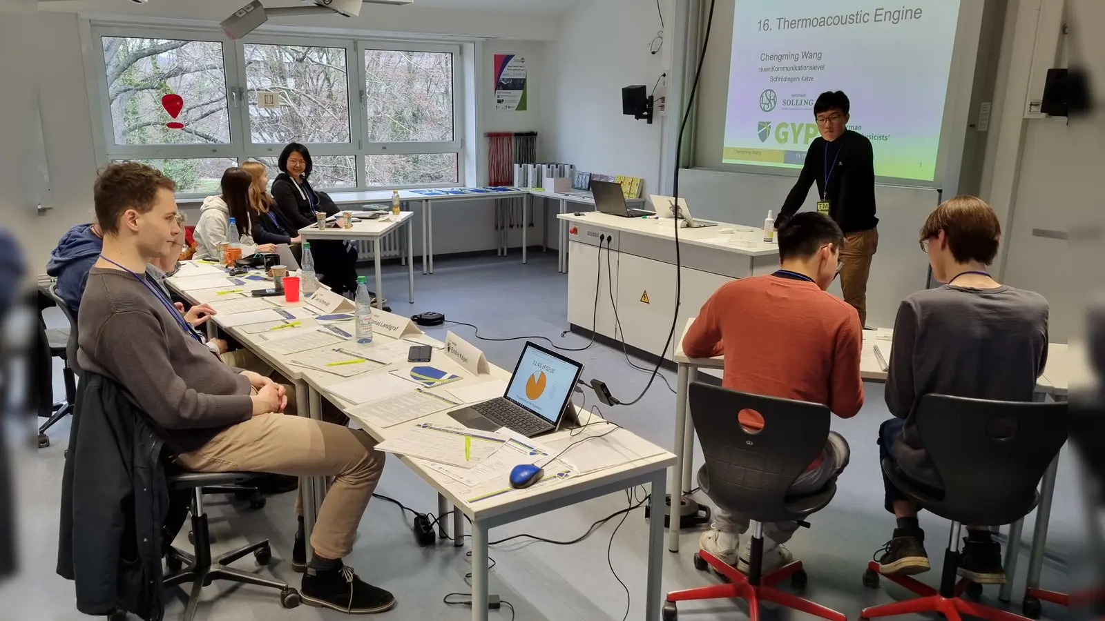 Beginn des „Physics Fight“ zwischen Chengming Wang vom Team „Kommunikationslevel Schrödingers Katze“ (hinterer Tisch) und Jeff Zeng vom Team „Schwarzschildkröten“ (rechter Tisch) vor den Augen der Jury (lange Tischreihe links). (Foto: privat)
